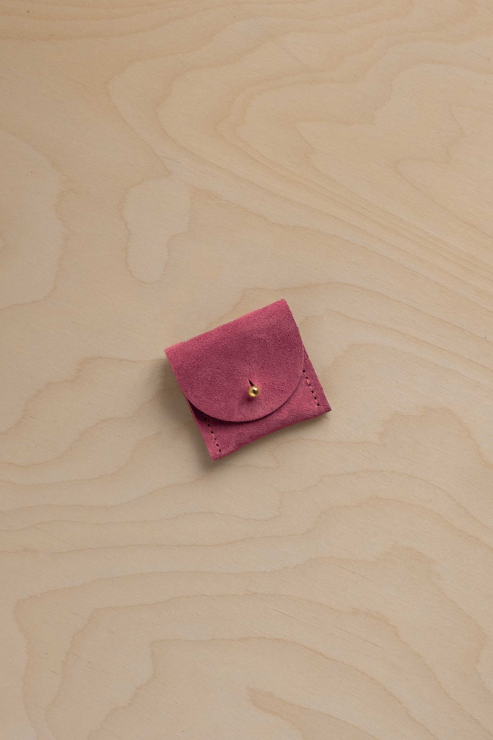 A small rose pink suede pouch with a gold button closure, placed on a light beige wood grain background.