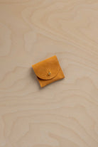 A small yellowy orange suede leather pouch with a flap closure and a gold button, placed on a light wood grain surface.