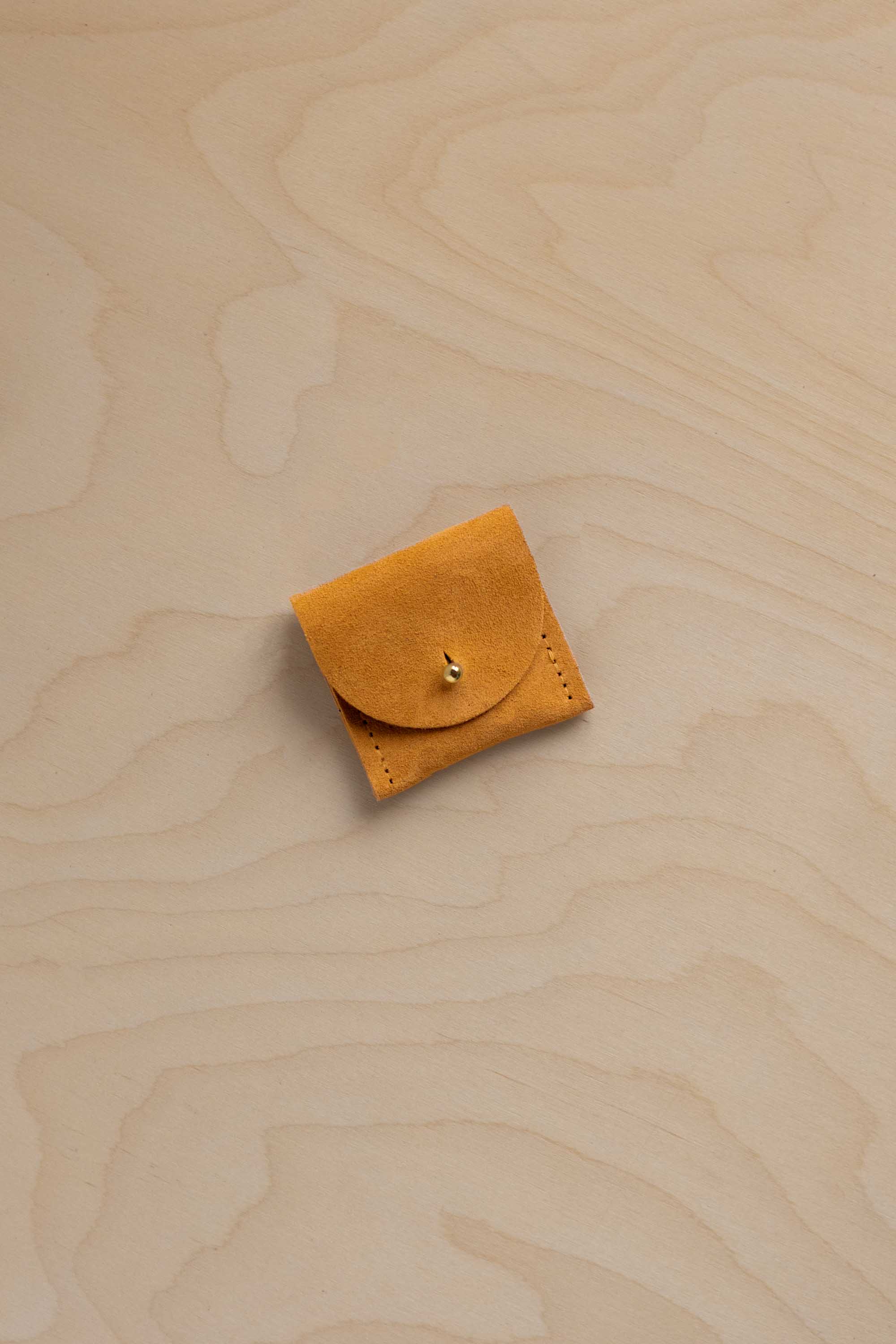 A small yellowy orange suede leather pouch with a flap closure and a gold button, placed on a light wood grain surface.