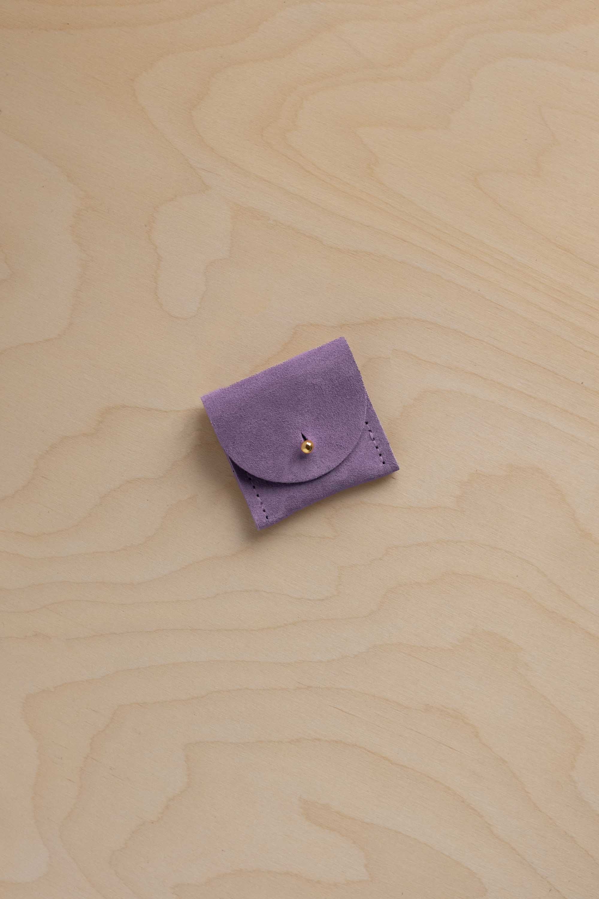 A small lavender purple suede ring pouch with a flap closure and a tiny gold button, placed on a light beige wood grain surface.