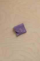 A small lavender purple suede ring pouch with a flap closure and a tiny gold button, placed on a light beige wood grain surface.