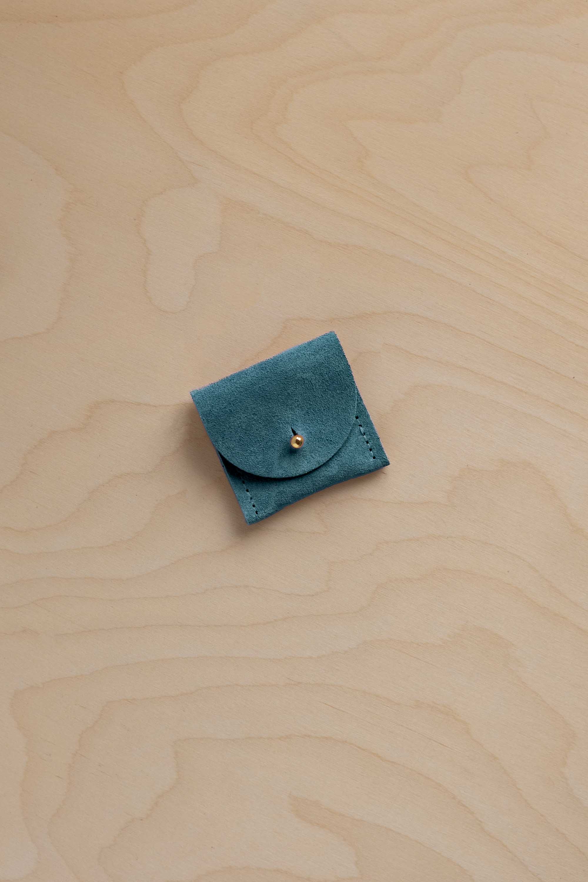 A small teal blue suede pouch with a flap closure and a tiny gold fastening, placed on a light wood grain surface.