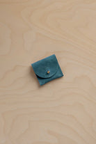 A small teal blue suede pouch with a flap closure and a tiny gold fastening, placed on a light wood grain surface.