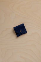 A small navy blue suede leather pouch with a flap closure and a gold-tone button, placed on a light wood grain surface.