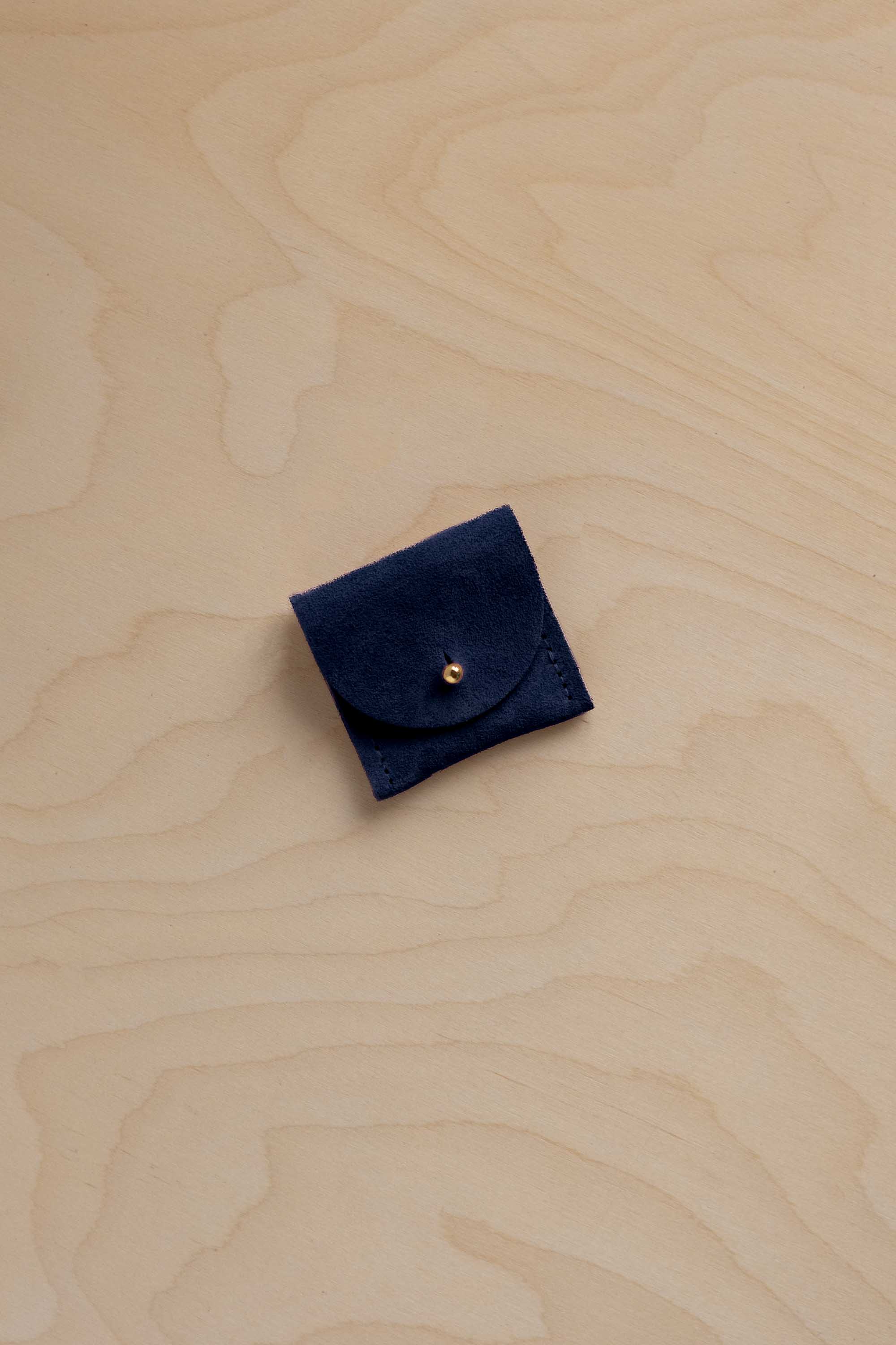 A small navy blue suede leather pouch with a flap closure and a gold-tone button, placed on a light wood grain surface.