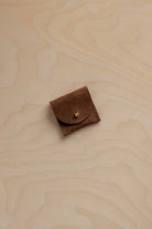 A small brown leather pouch with a flap closure and a gold-tone snap button, placed on a light wood grain surface.