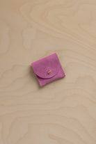 A small light magenta pink suede leather ring pouch with a flap closure and a tiny gold button, placed on a light wood grain surface.