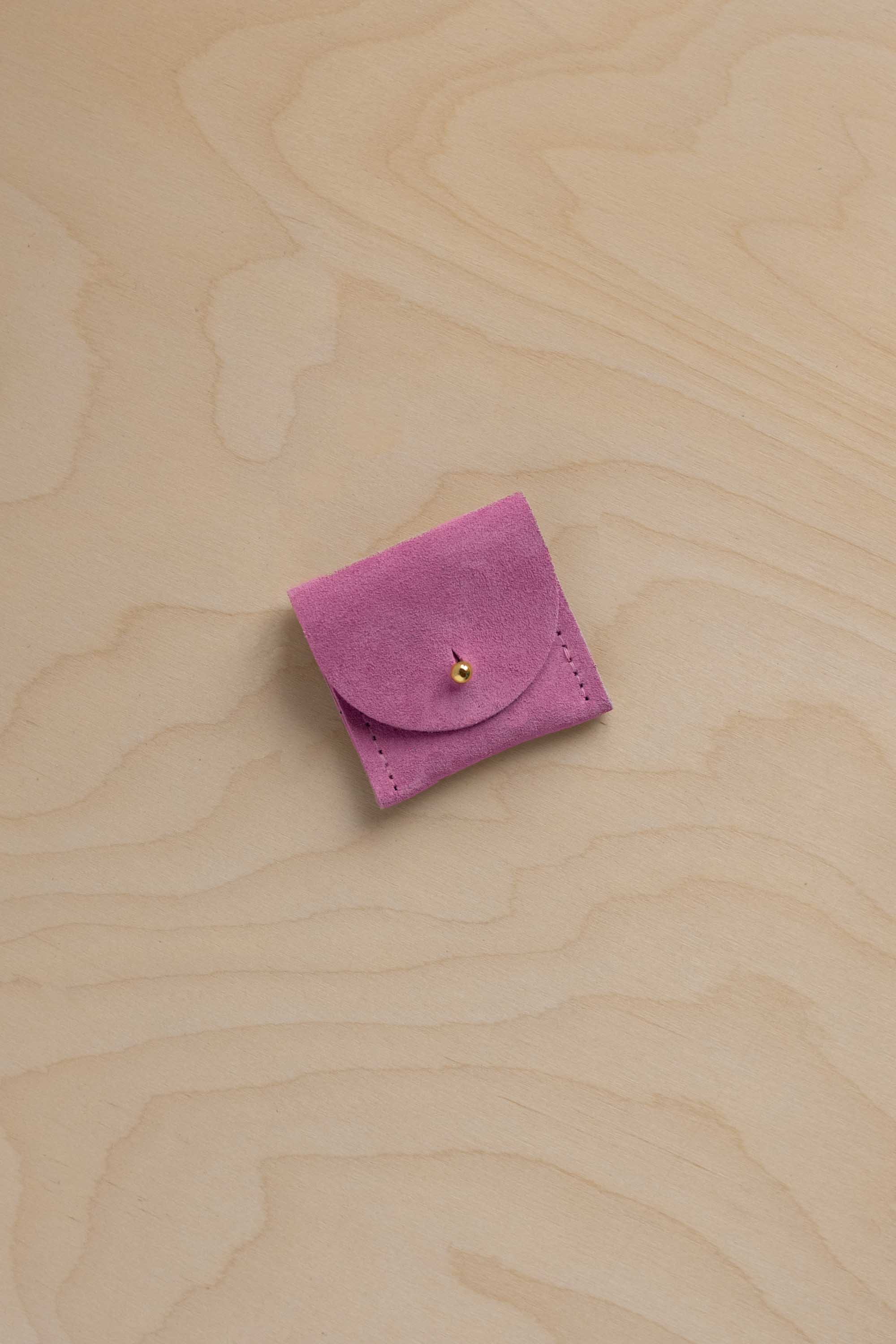 A small light magenta pink suede leather ring pouch with a flap closure and a tiny gold button, placed on a light wood grain surface.