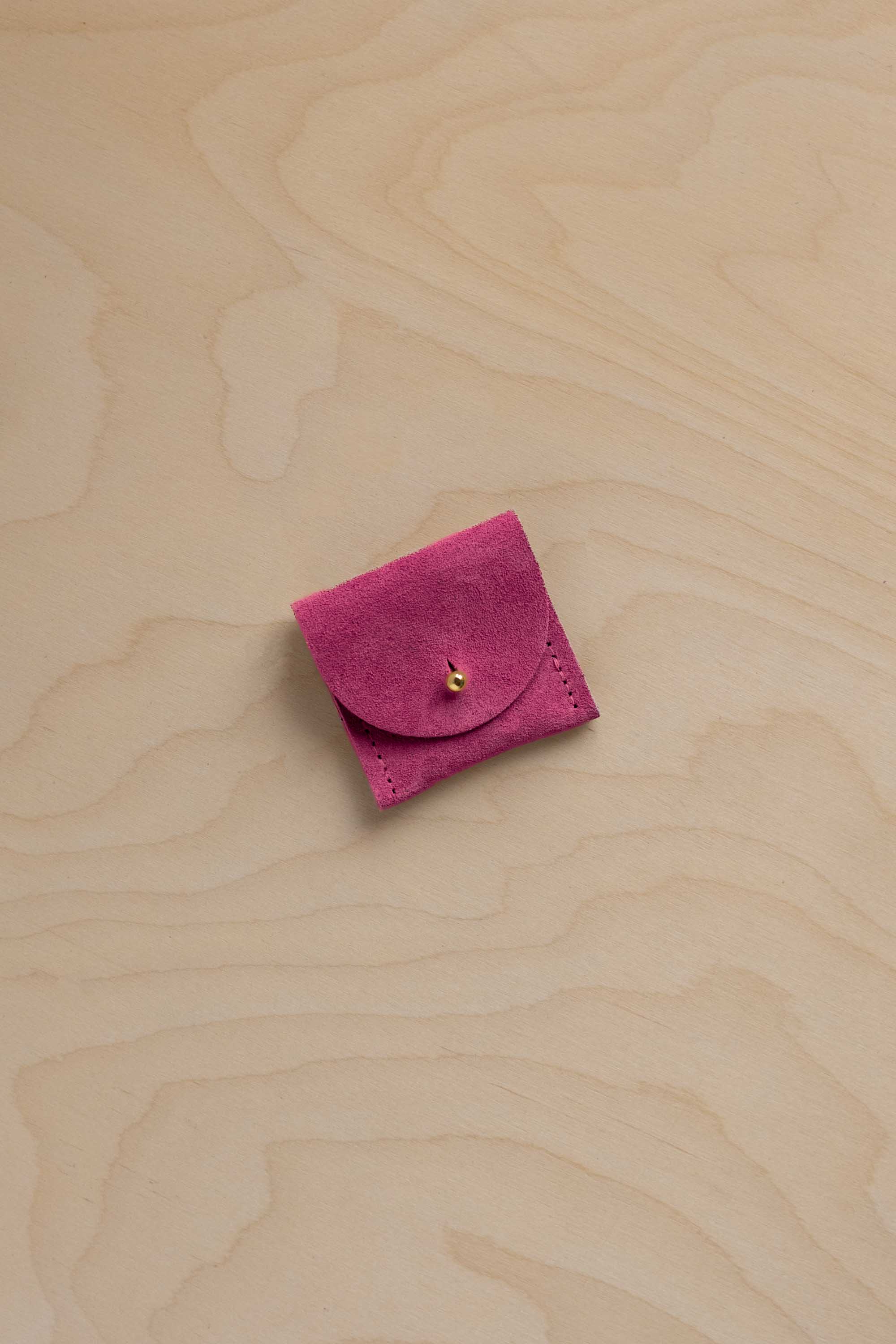 A small magenta pink suede jewelry pouch with a flap closure and a tiny gold bead accent, placed on a light wood grain background.