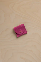 A small magenta pink suede jewelry pouch with a flap closure and a tiny gold bead accent, placed on a light wood grain background.