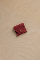 A small rust red suede pouch with a gold button closure, placed on a light wood grain background.
