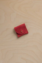 A small red suede jewellery pouch with a circular flap and a tiny gold button, placed on a light wood grain surface.