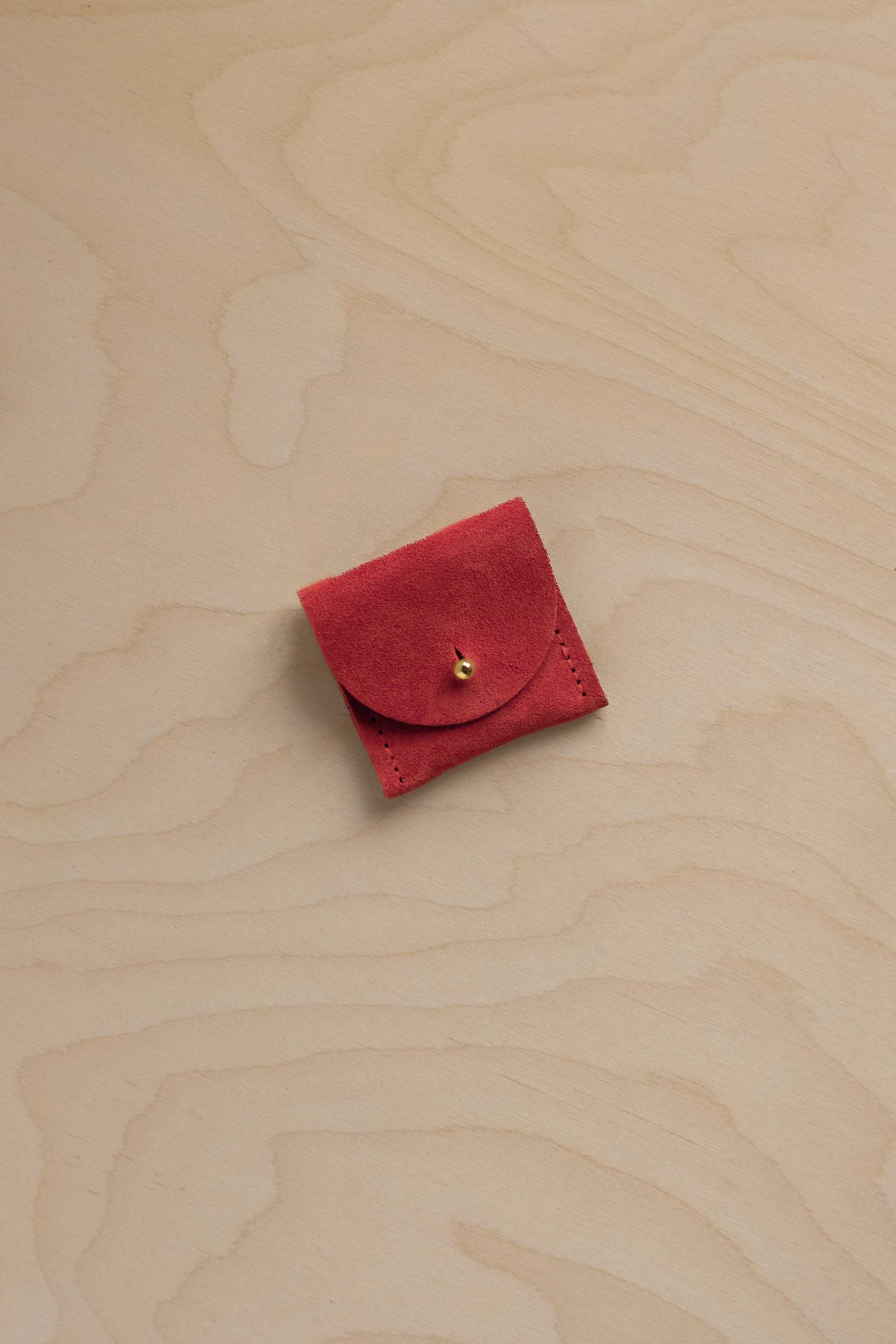 A small red suede jewellery pouch with a circular flap and a tiny gold button, placed on a light wood grain surface.