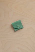 A small light emerald green suede pouch with a flap closure and a tiny gold button, placed on a light beige wood-grain surface.