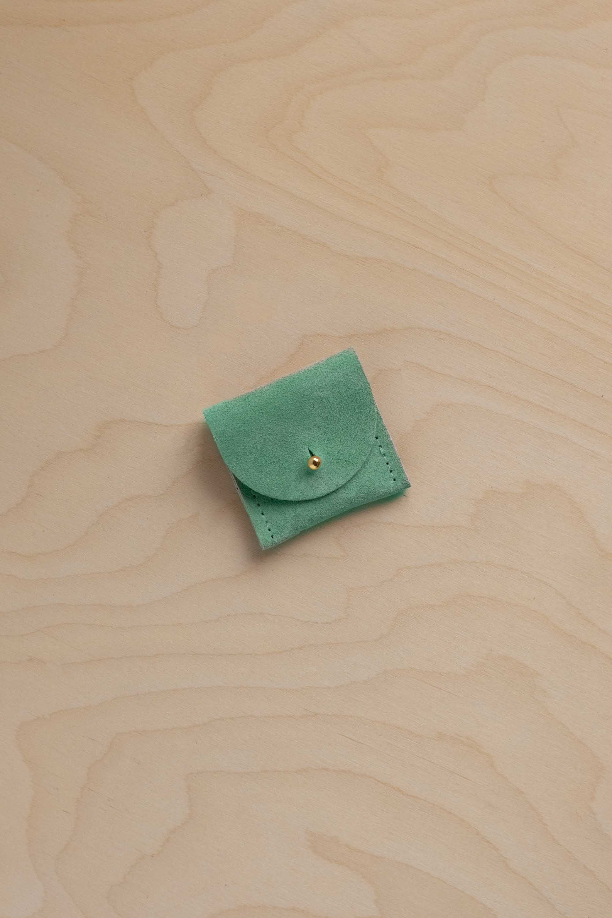 A small light emerald green suede pouch with a flap closure and a tiny gold button, placed on a light beige wood-grain surface.