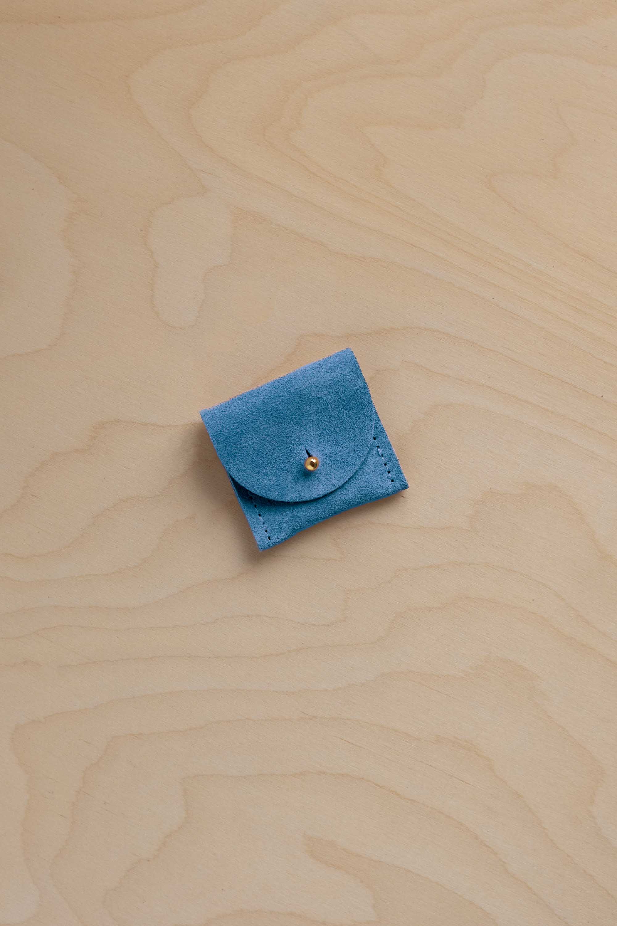 A small bright blue suede leather jewellery pouch with a flap closure and a tiny gold bead accent, placed on a light wood grain surface.