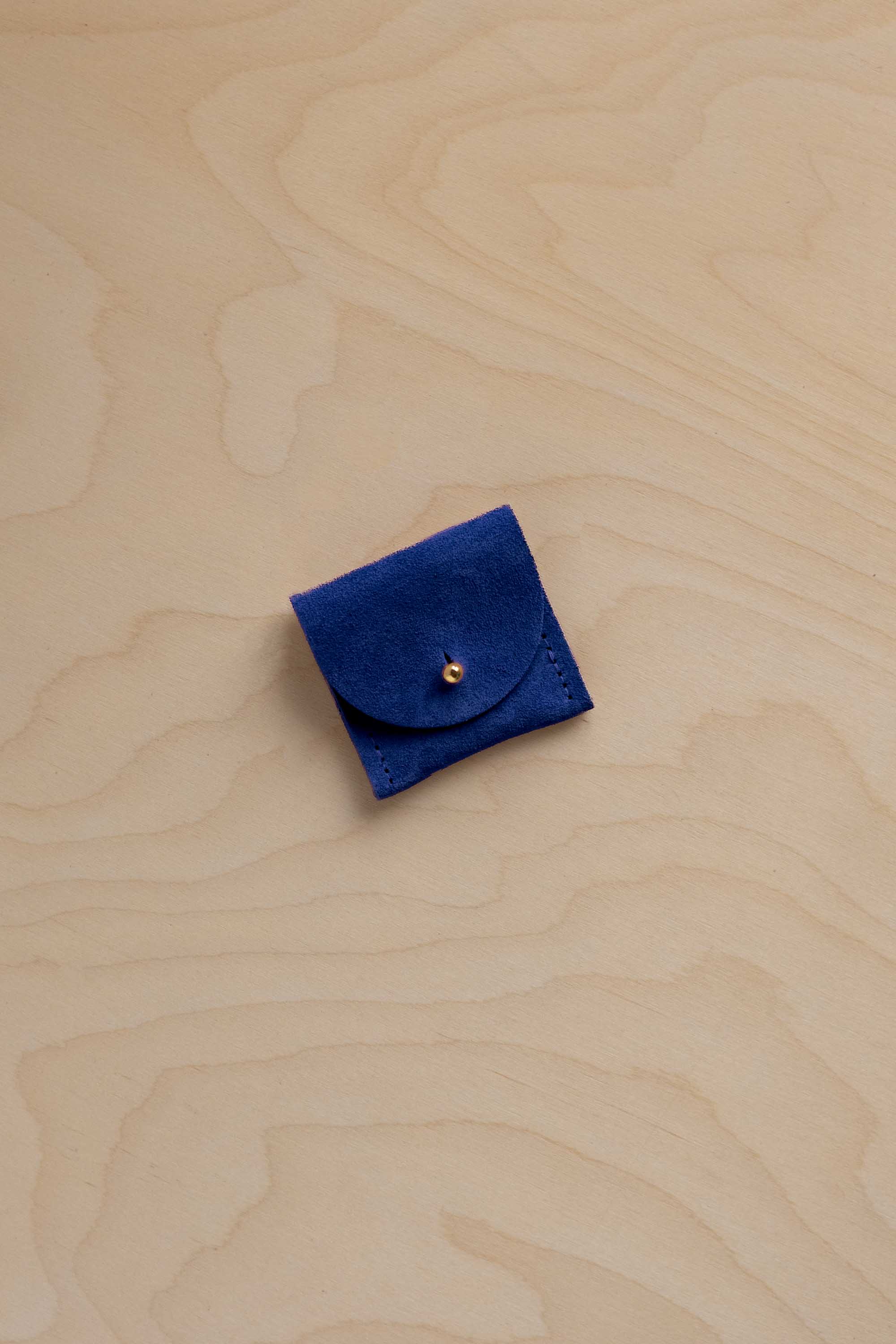 A small square dark blue suede pouch with a flap closure and a single gold button, placed on a light wood grain surface.