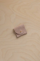 A small dusty beige / pink suede jewelry pouch with a gold button, placed on a light wood grain background.
