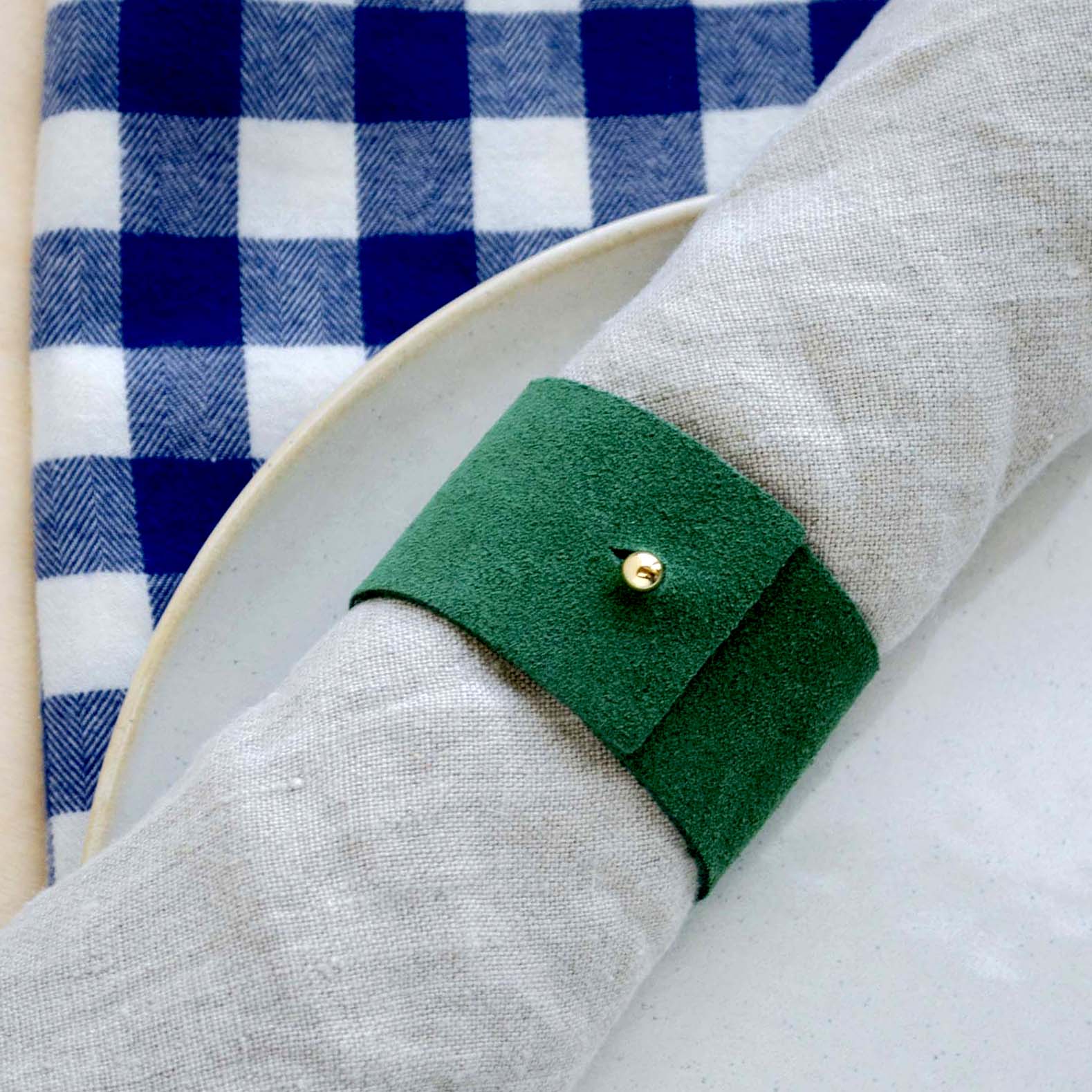Moss green suede napkin ring with gold stud on a stoneware plate and a blue gingham table cloth for Christmas.