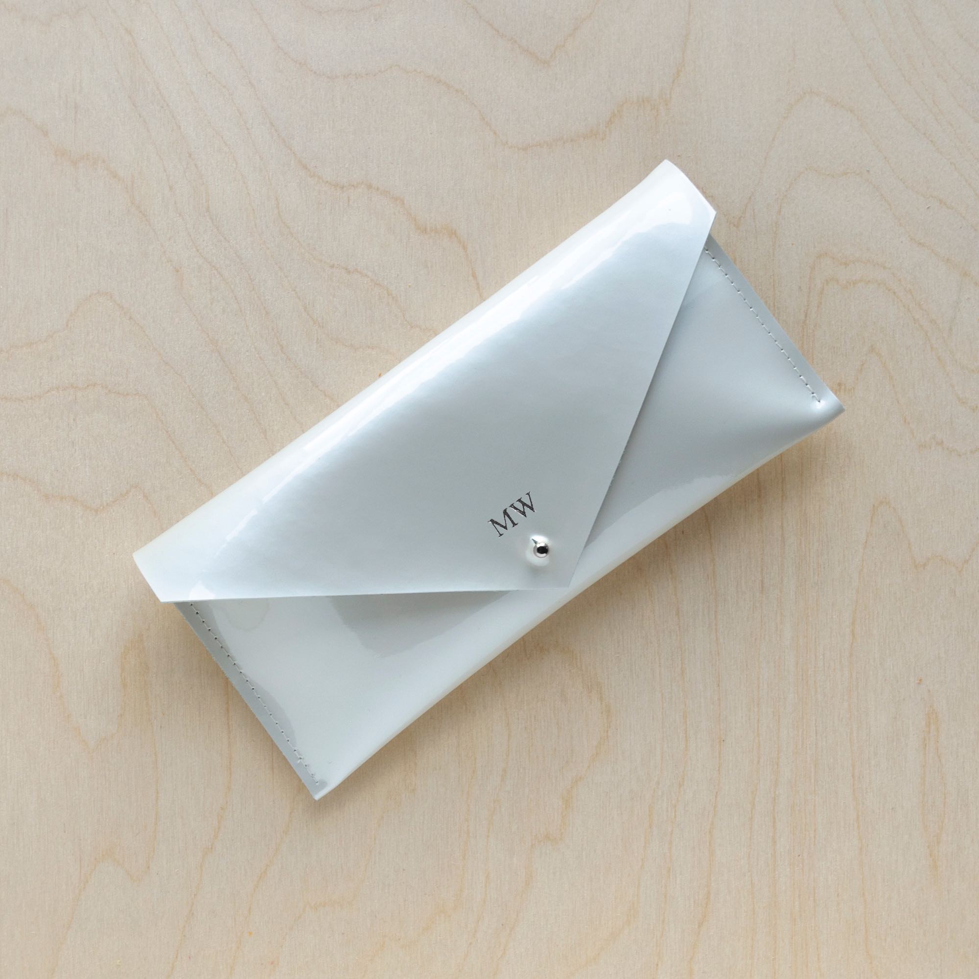 A white pearlescent leather clutch purse with a minimalist envelope design, displayed against a wooden background.