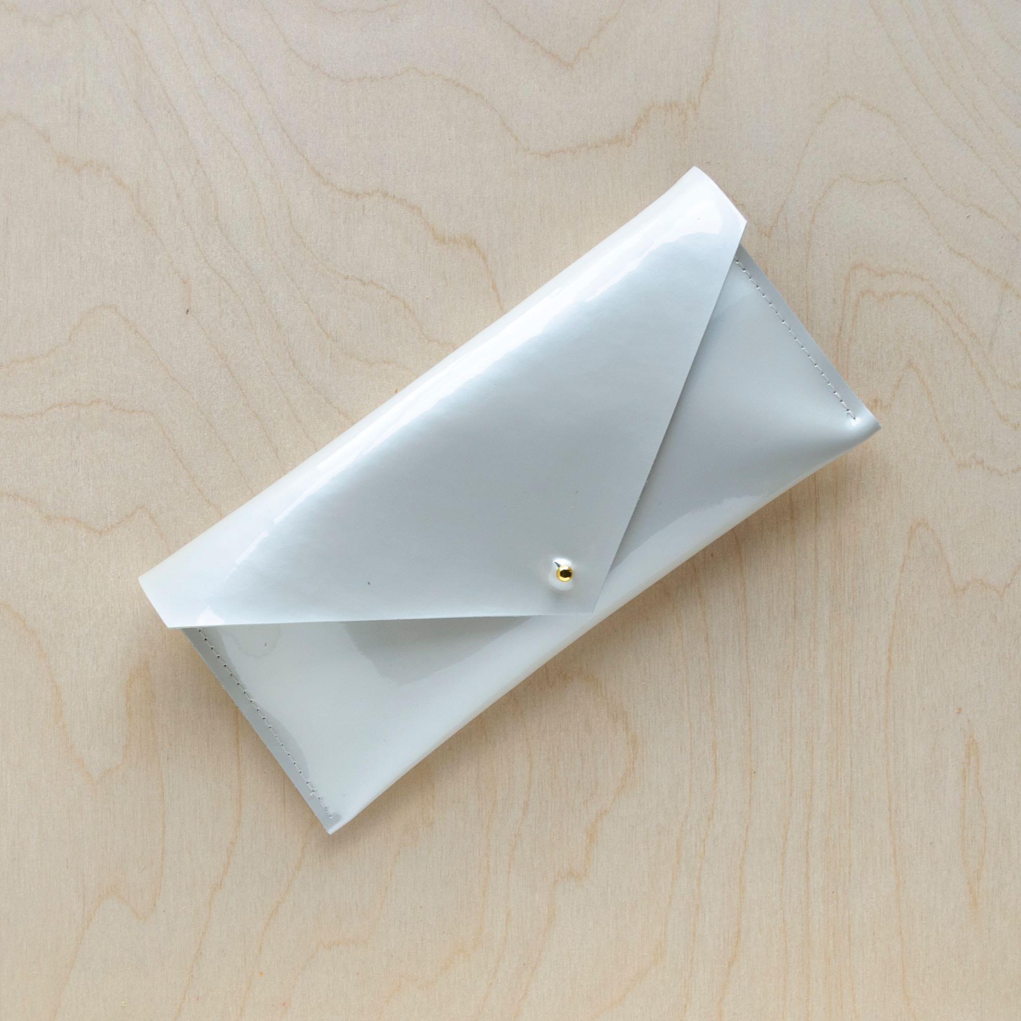 Envelope style compact clutch bag in a mother of pearl leather sits against a wooden background with gold hardware.