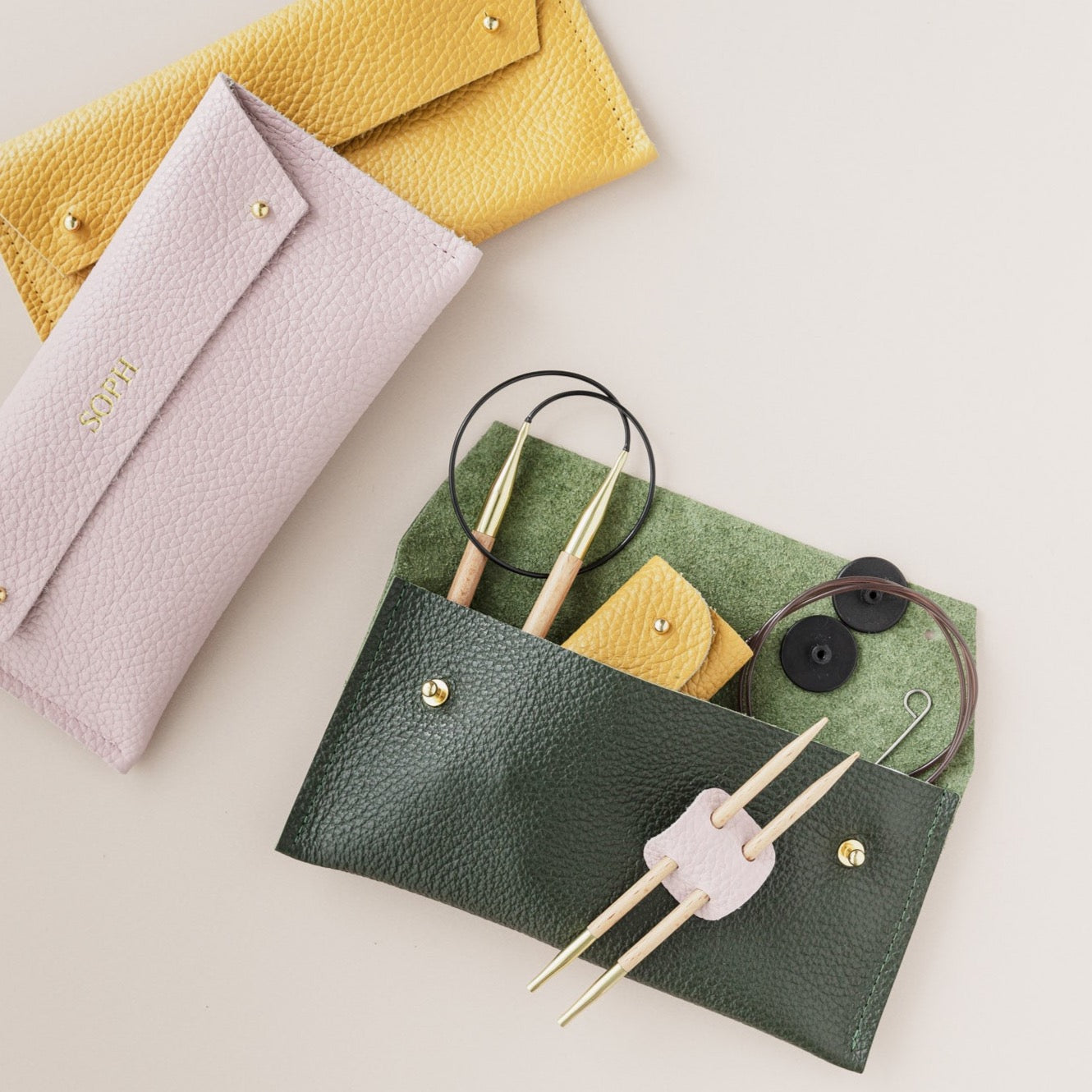 Yellow, pink and green sewing cases sit on a cream background. The green pouch shows circular knitting needles and notions inside it.