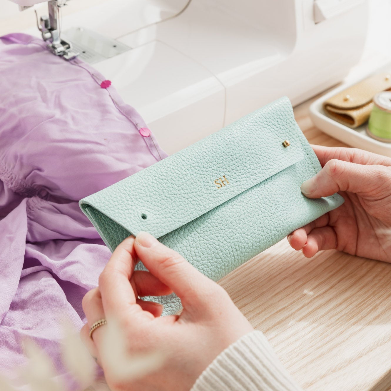 Sewer holds light turquoise leather sewing case with two metal studs while she sews lilac dress. The leather pouch is personalised with the initials SH.