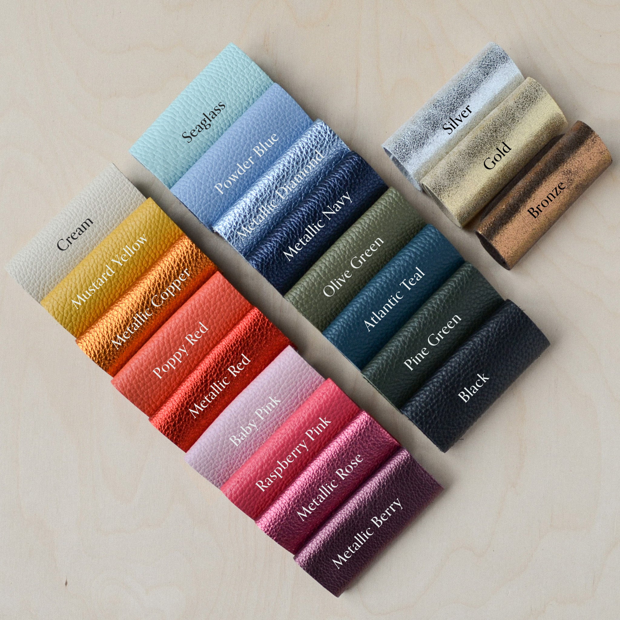 A set of Italian leather color swatches arranged on a light wood background, showing various colors and metallic finishes including Cream, Mustard Yellow, Powder Blue, Metallic Navy, Silver, Gold, Bronze, Olive Green, Atlantic Teal, Pine Green, Black, Metallic Rose, and Metallic Berry.