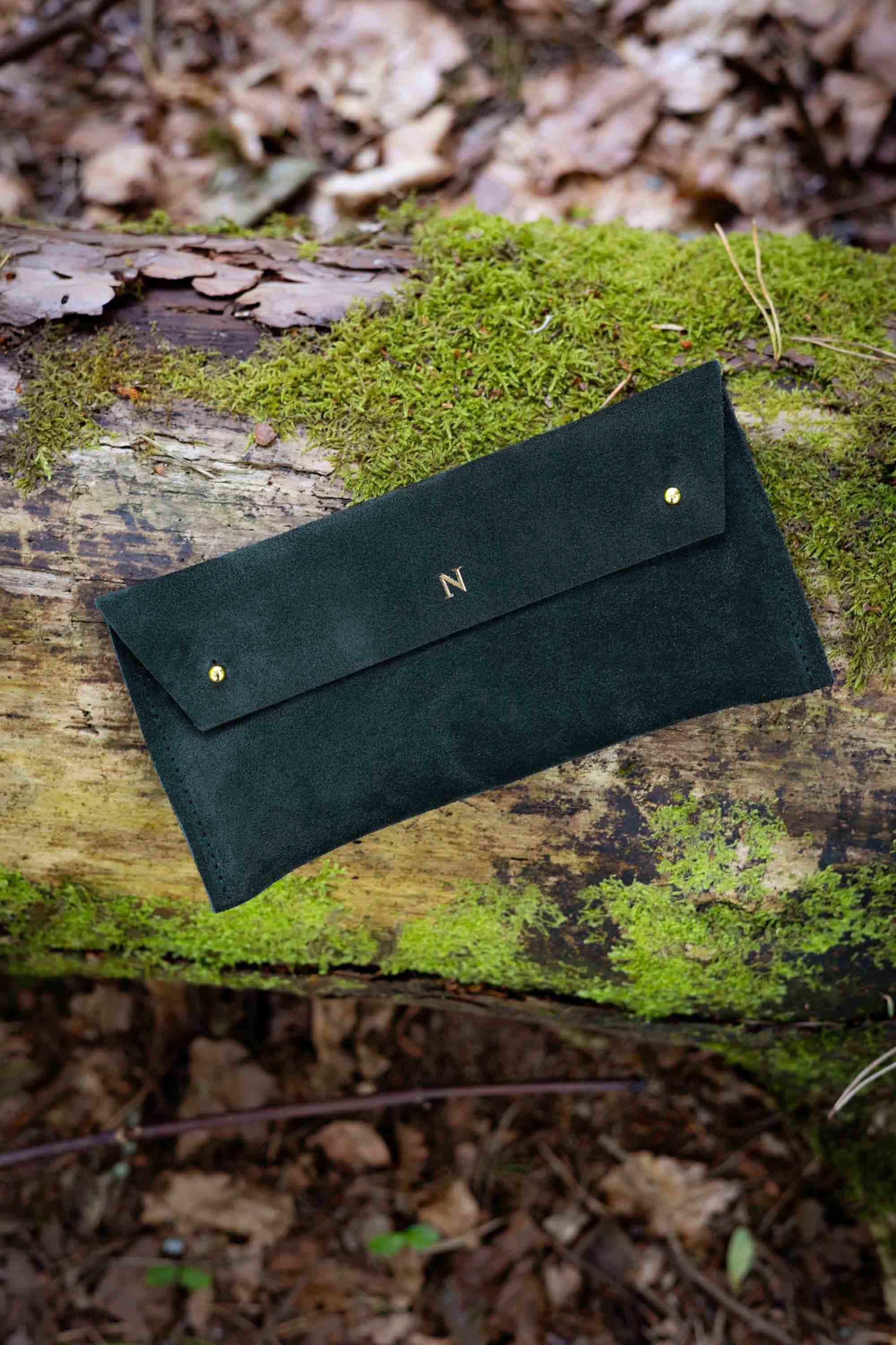 A dark forest green suede clutch or wallet with gold-tone studs and a small monogram 'N' on the flap, resting on a mossy log in a forest setting.