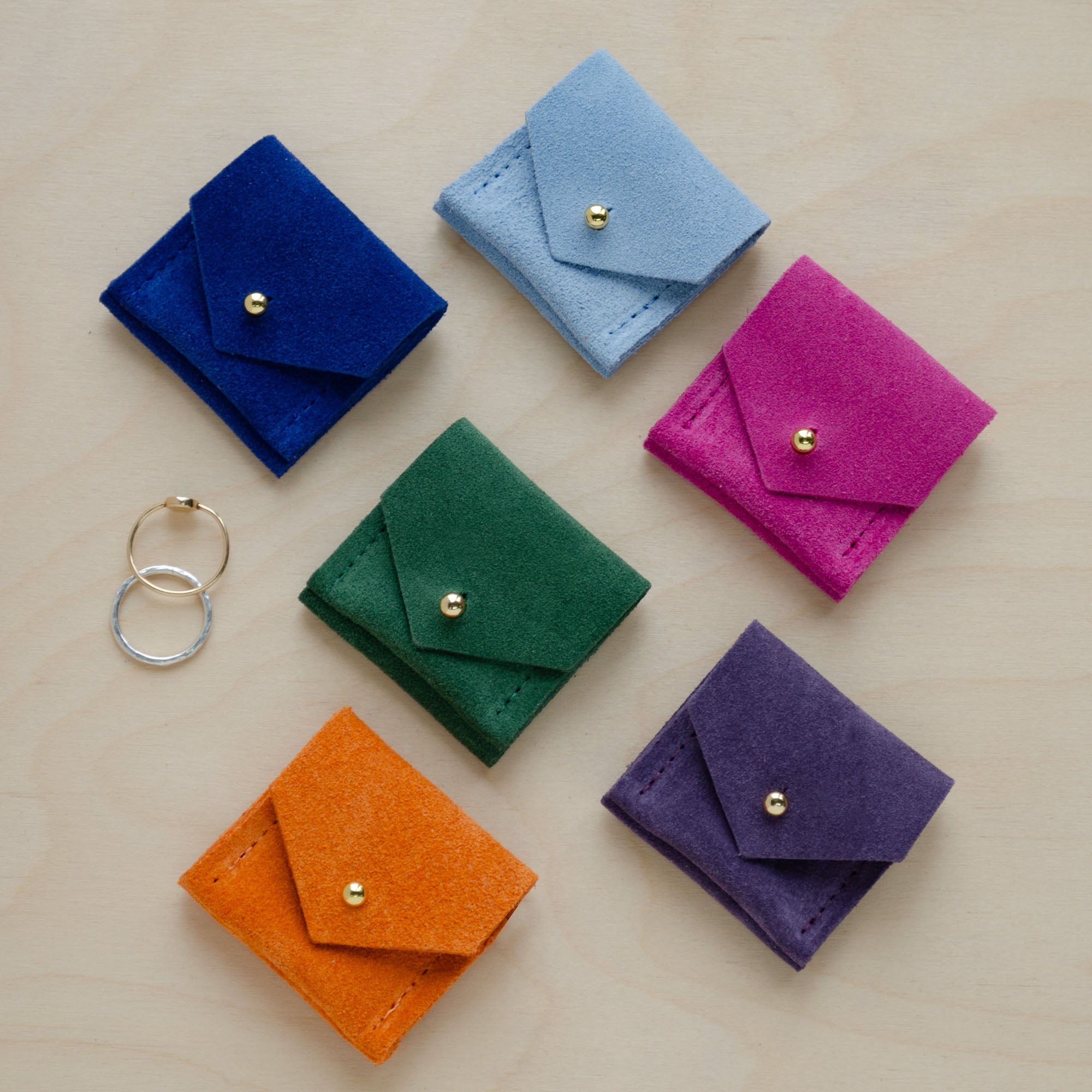 Six of the Wedding Ring Pouches in suede. The colours are, Ultramarine Blue, Pale Blue, Moss Green, Fuchsia Pink, Clementine Orange and Grape Purple.