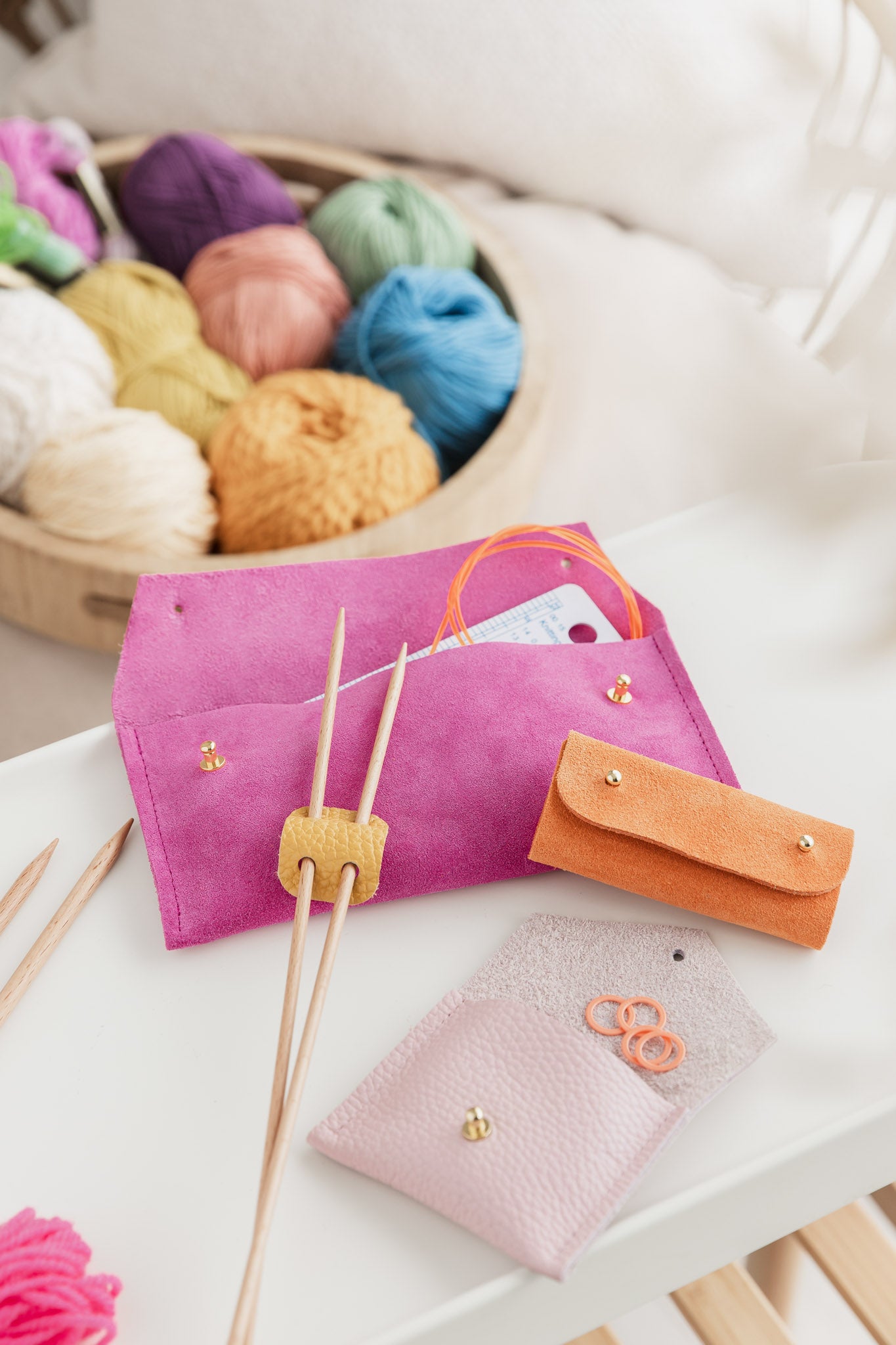 Knitting needle holder in yellow leather sits on pink notion pouches and orange suede sewing needle case. In the background there is a basket of colourful wool.