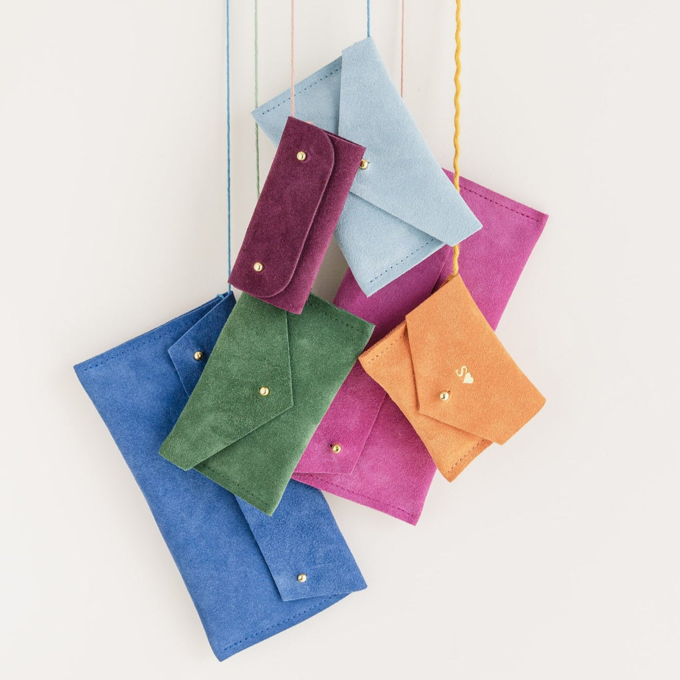 A group of pouches hanging from thread, the colours include: Plum, Pale Blue, Moss Green, Orange, Ultramarine and Fuchsia pink. 