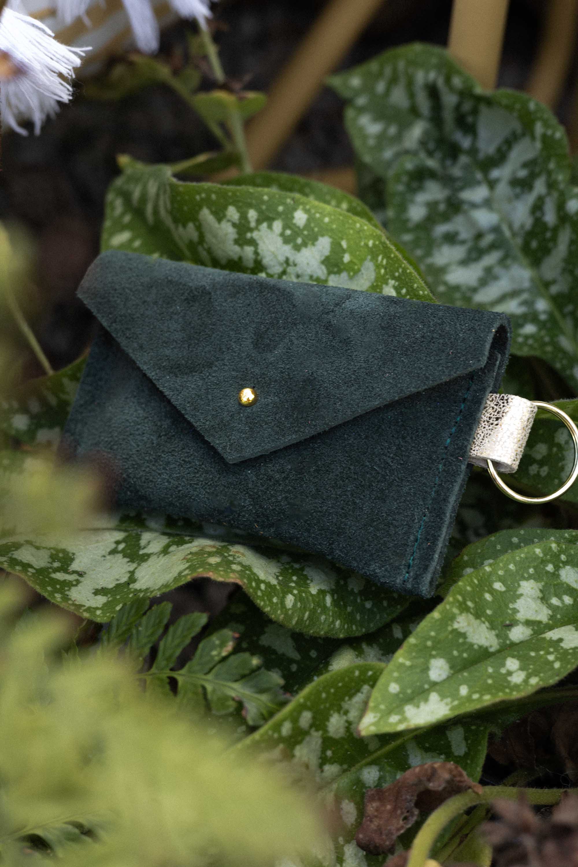 A small deep forest green coloured suede envelope-style keyring pouch with a gold button, nestled amongst some leaves in nature.