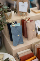 A photo of a retail display featuring suede pouches in different styles and assorted colours on a light wood grain shop display, with a small plant and a hanging display of leather earrings.