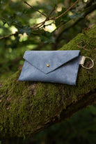 A small dusky blue coloured suede envelope-style keyring pouch with a gold button, resting on a tree branch in a forest.
