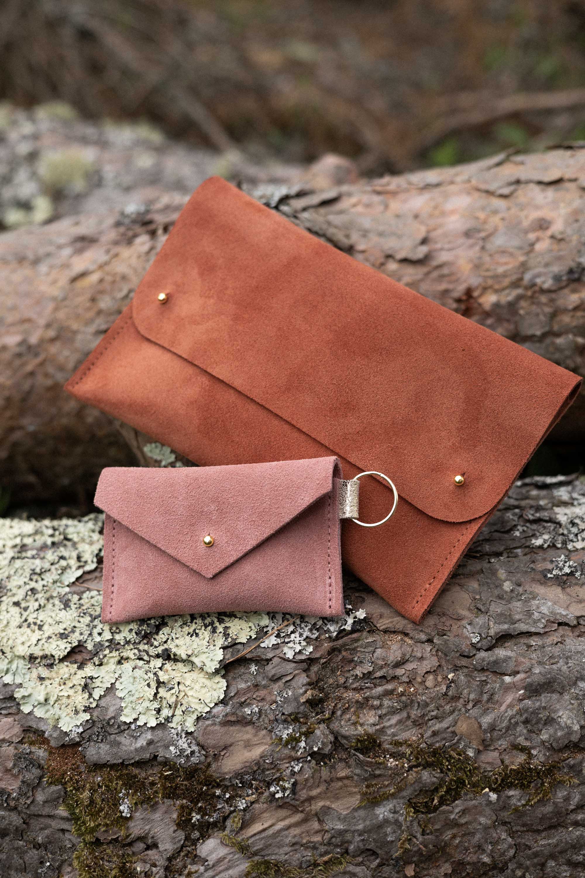 A small dusky rose coloured suede envelope-style keyring pouch with a gold button, resting on a light wood grain surface, alongside a rust coloured suede clutch bag.