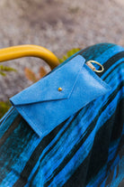 A small sky blue coloured suede envelope-style keyring pouch with a gold button, resting on a blue and black striped  patterned fabric.