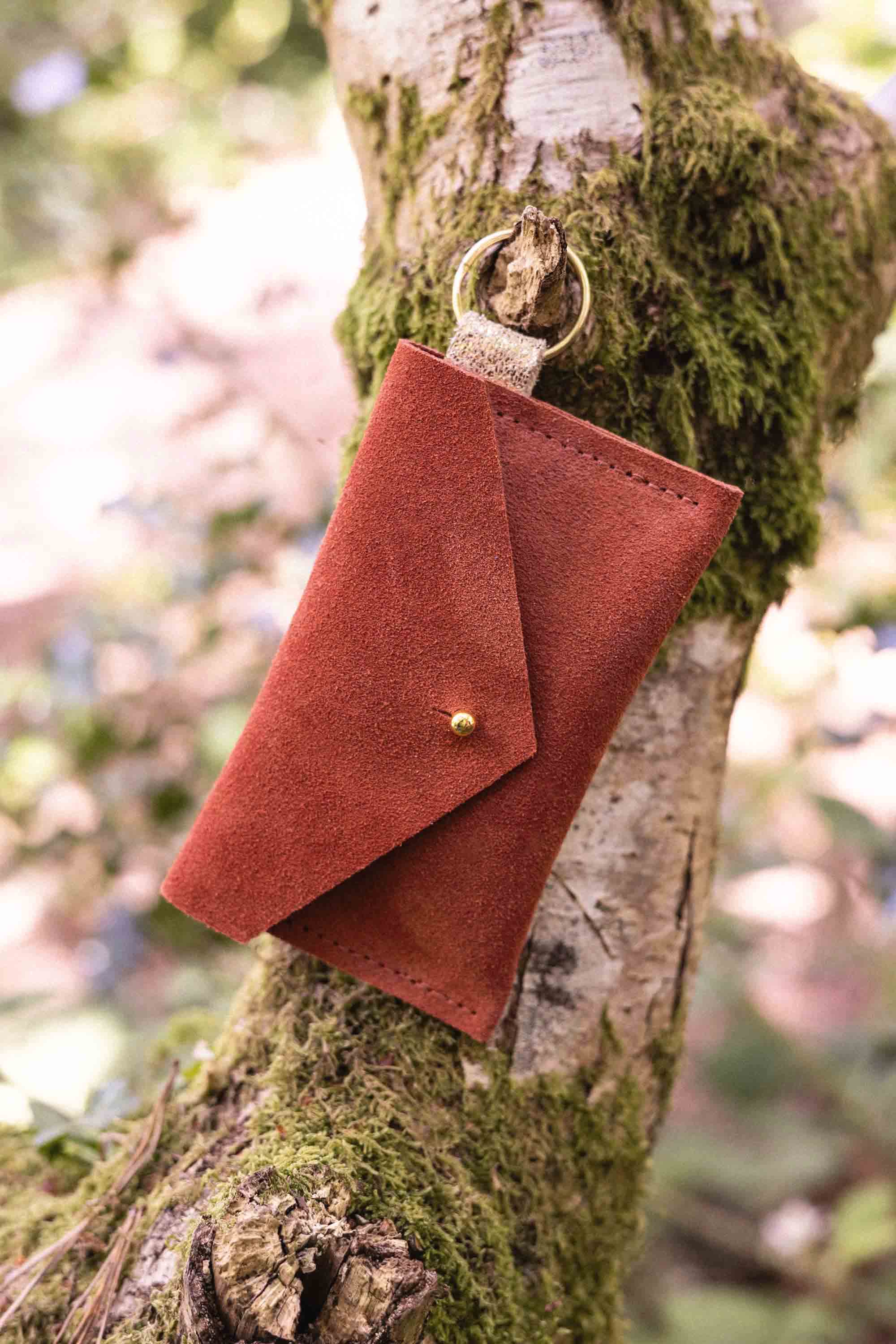A small terracotta coloured suede envelope-style keyring pouch with a gold button, hanging from a branch with a blurred background.
