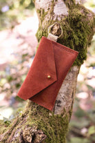 A small terracotta coloured suede envelope-style keyring pouch with a gold button, hanging from a branch with a blurred background.