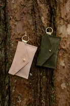 Two small suede envelope-style keyring pouches with a gold button, resting against a textured tree.