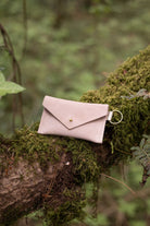 A small warm sand coloured suede envelope-style keyring pouch with a gold button, resting on a branch in a forest.