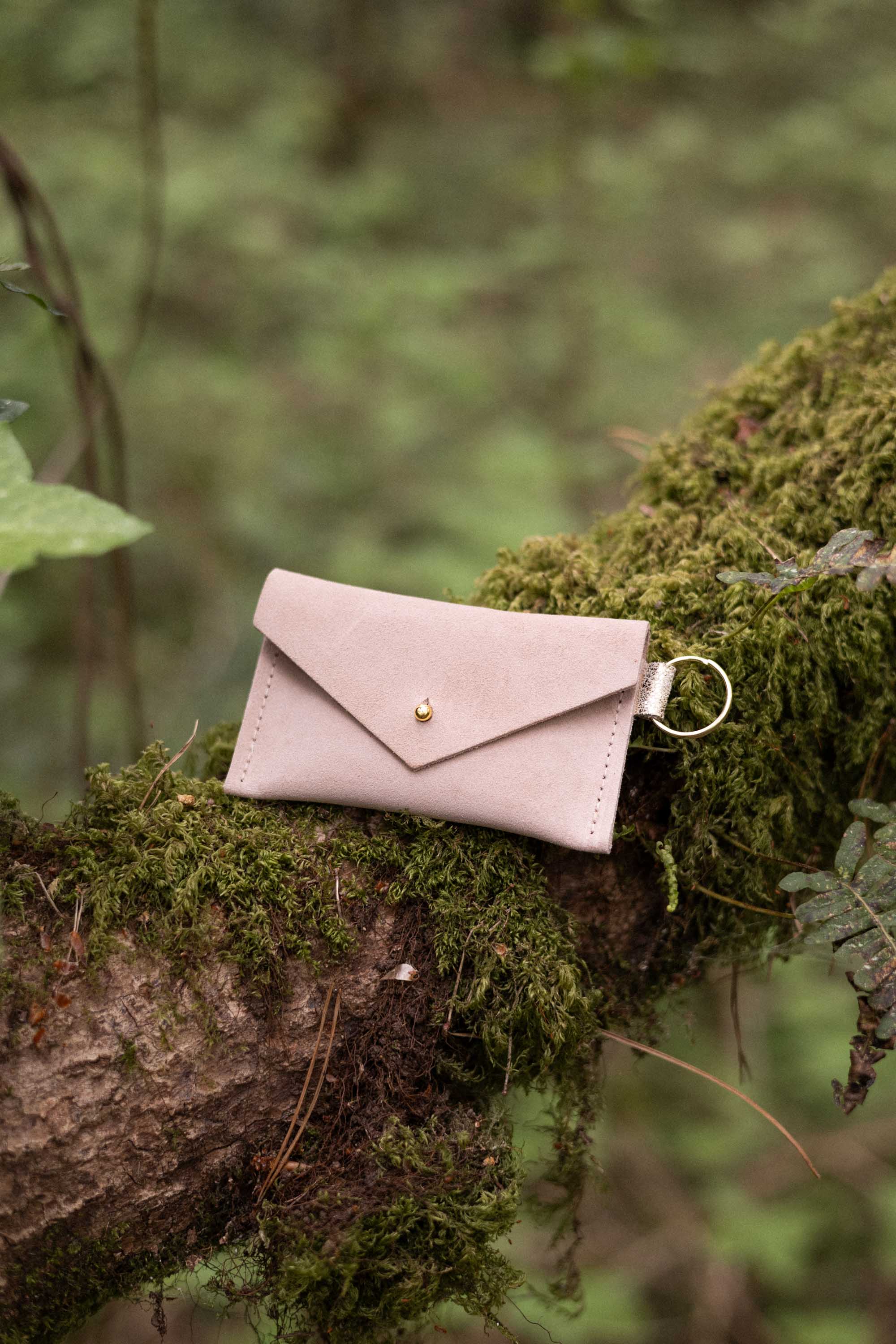 A small warm sand coloured suede envelope-style keyring pouch with a gold button, resting on a branch in a forest.