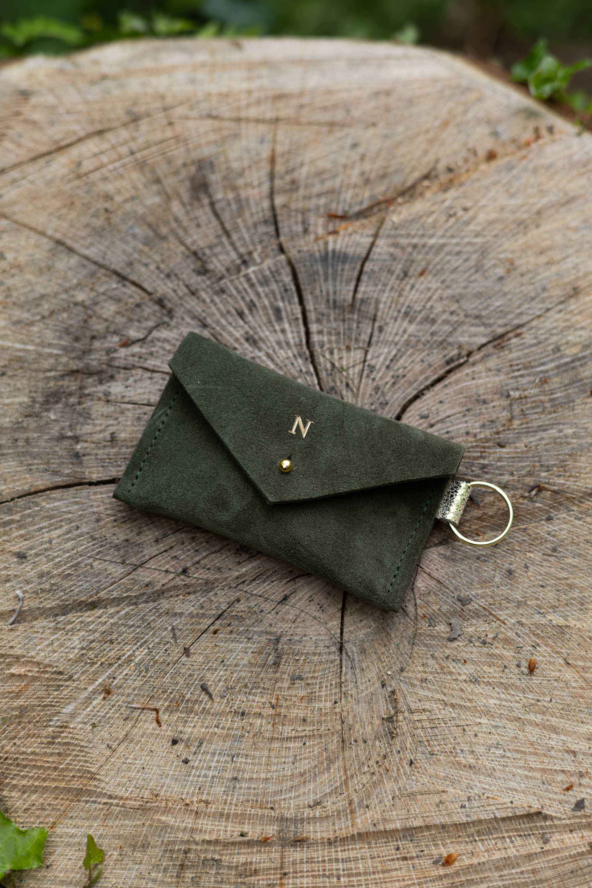 Small soft deep forest coloured suede keyring pouch with a gold foiled initial on the front, resting against a tree.