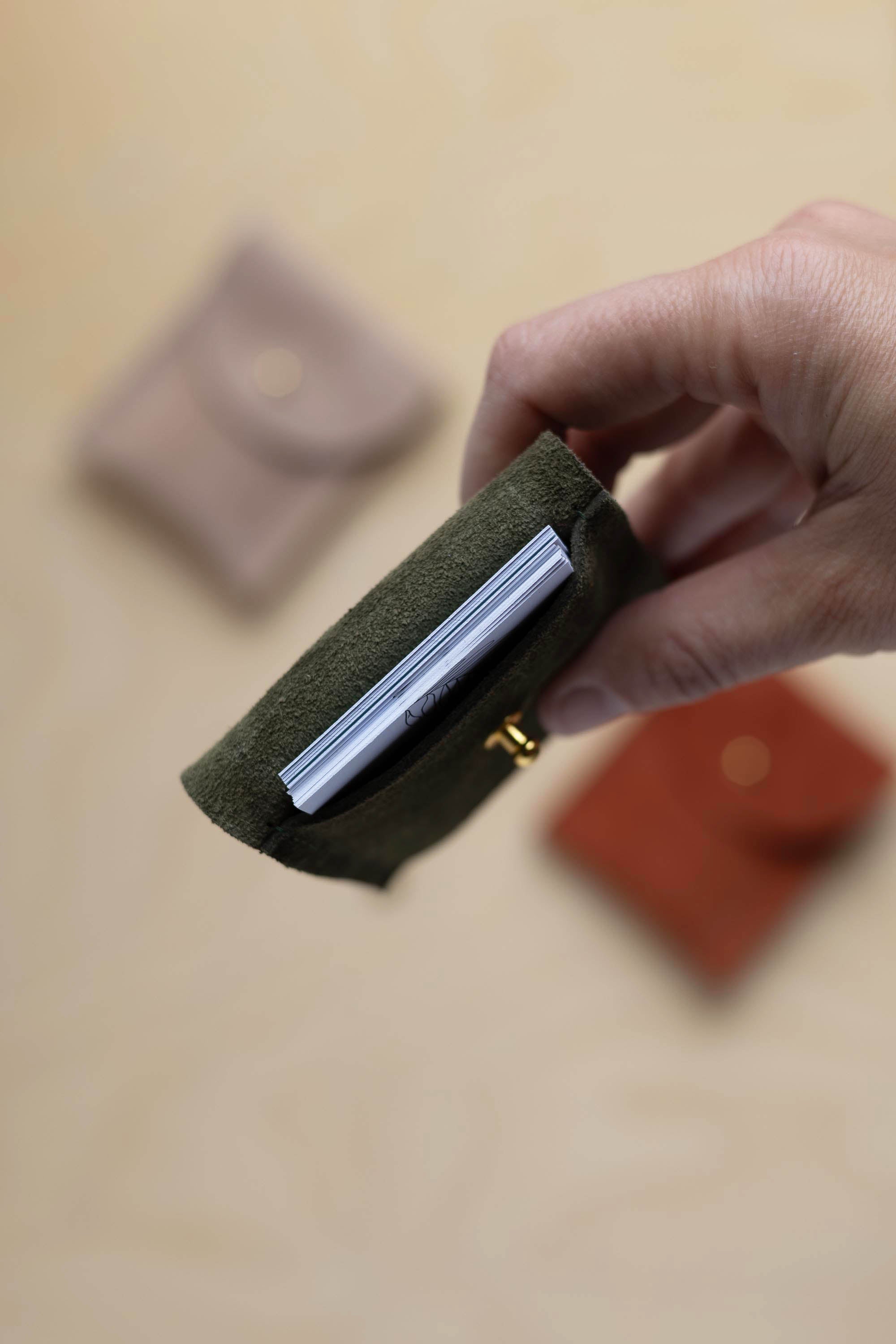 A close up view of a hand holding a deep forest coloured suede pouch with a gold coloured fastening with business cards nestled inside. In the background there are two coloured suede pouches on a light wood grain surface.