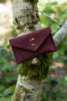 A small burgundy suede pouch with gold foiled initial and heart shape on the front, with a flap and gold button fastening, leaning against a tree branch with an outdoor background.