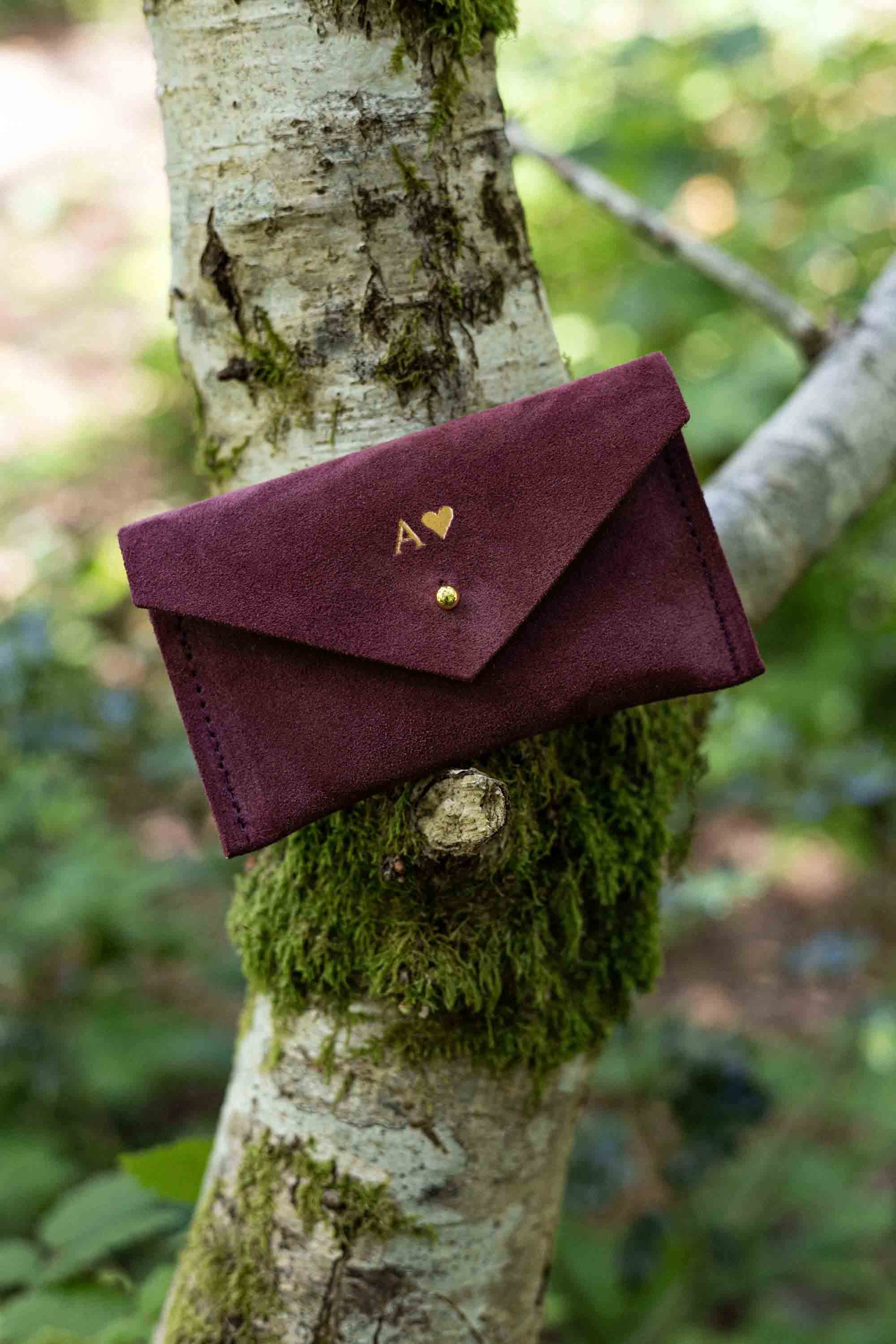 A small burgundy suede pouch with gold foiled initial and heart shape on the front, with a flap and gold button fastening, leaning against a tree branch with an outdoor background.