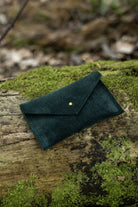 A small deep forest green coloured suede pouch with a flap closure and gold coloured fastening, with an outdoor background.