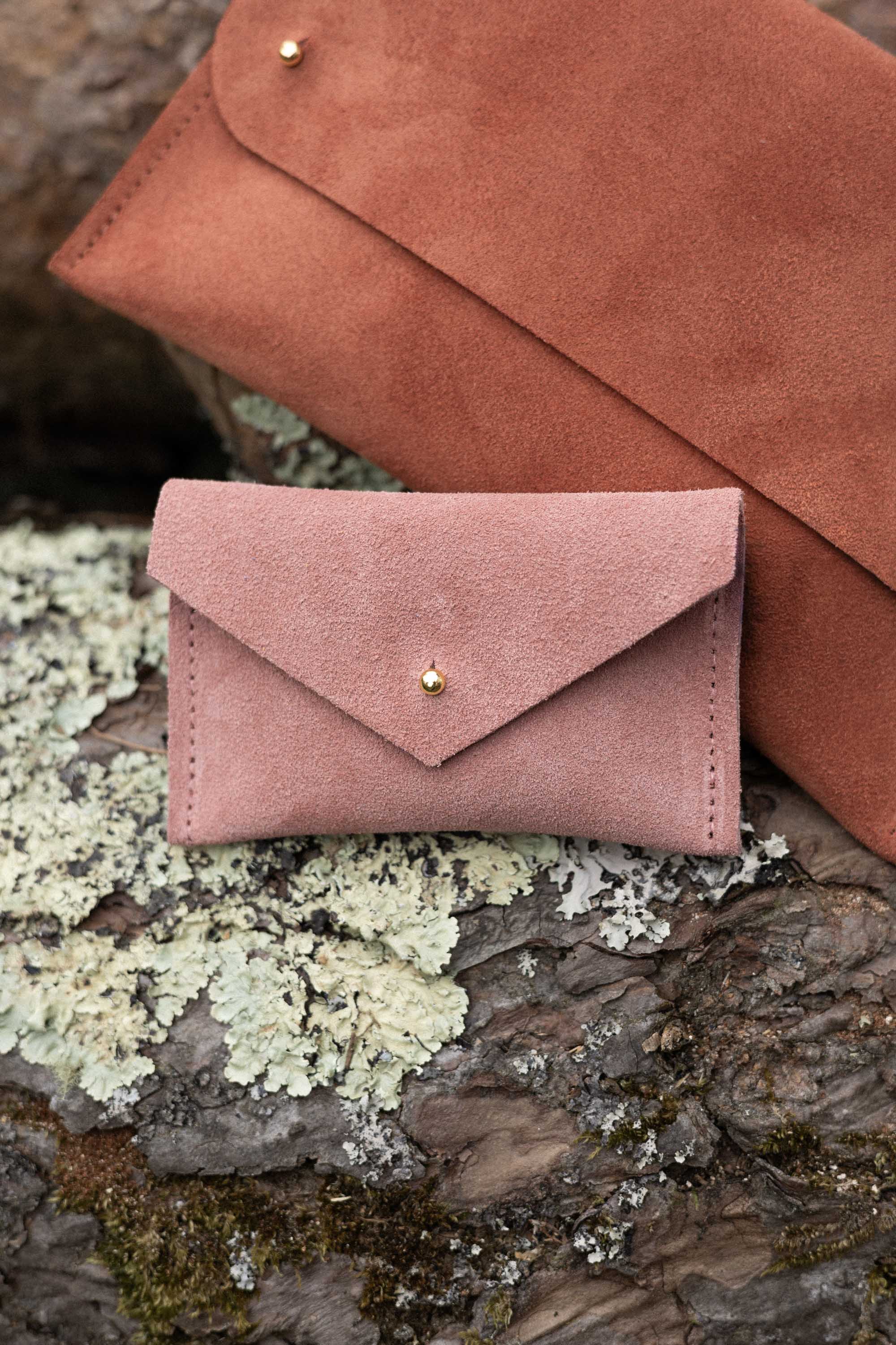 A small dusty rose coloured suede pouch with a flap closure and gold coloured fastening, on a natural wood surface, alongside a terracotta suede clutch bag.