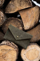 A small soft olive coloured suede pouch with a flap closure and gold coloured fastening, leaning against a wooden natural surface.