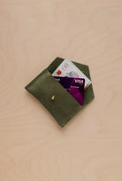 A small soft olive suede pouch with a bank card and bank note nestled inside with a flap and gold button fastening, on a light wood grain surface.